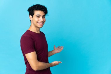 Genç Venezuelalı bir adam mavi arka planda izole edilmiş. Davet için ellerini yana uzatıyor.