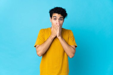 Genç Venezuelalı adam mavi arka planda izole edilmiş, ağzı elleriyle kapatıyor.