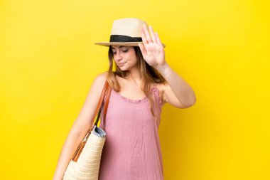 Sarı arka planda izole edilmiş plaj çantası tutan beyaz bir kadın dur hareketi yapıyor ve hayal kırıklığına uğruyor.