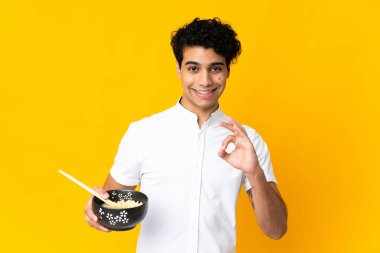 Genç Venezuelalı bir adam sarı arka planda izole edilmiş. Elinde yemek çubuklarıyla bir kase erişte tutarken parmaklarıyla işaret veriyor.