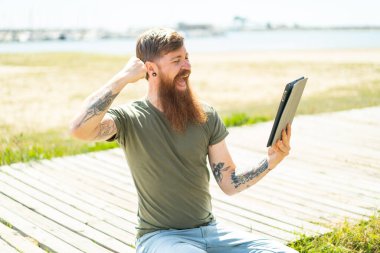 Kızıl saçlı, sakallı bir adam dışarıda bir tablet tutuyor ve zaferi kutluyor.