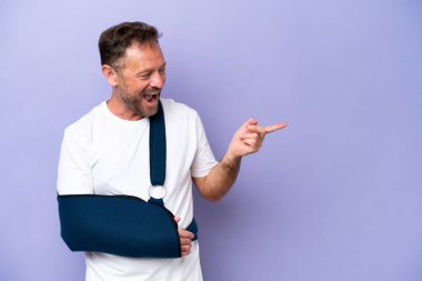 Kırık kollu, mor arka planda askısı olan orta yaşlı, beyaz bir adam.