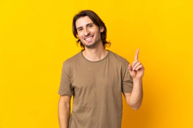 Sarı arka planda izole edilmiş genç yakışıklı adam en iyinin işareti olarak parmağını kaldırıyor.