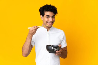 Genç Venezuelalı adam sarı arka planda izole edilmiş. Elinde çubuklu erişte kasesi var.