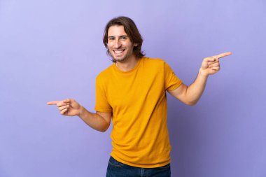 Mor arka planda izole edilmiş genç yakışıklı adam parmak uçlarını işaret ediyor ve mutlu.