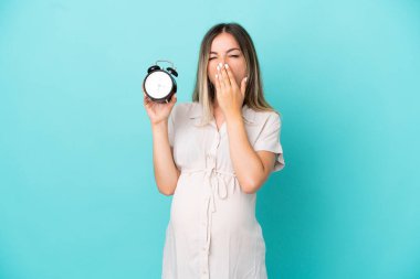 Genç Romen kadın mavi arka planda izole edilmiş hamile ve esnerken saati tutuyor