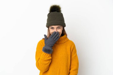 Kışlık şapkalı genç kız beyaz arka planda izole edilmiş mutlu ve gülümseyen ağızla elini kapatıyor.