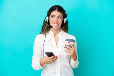 Tele pazarlamacı İtalyan bir kadın mavi arka planda izole edilmiş bir kulaklıkla çalışıyor. Elinde kahve ve bir şeyler düşünürken cep telefonu var.