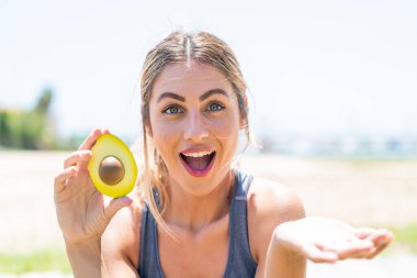 Sarışın bir kadın açık havada elinde bir avokado tutuyor ve yüzünde şok ifadesi var.