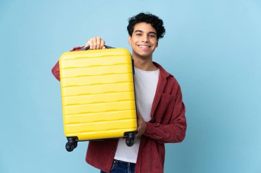 Genç Venezuelalı adam mavi arka planda izole edilmiş seyahat valiziyle.