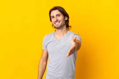 Sarı arka planda izole edilmiş genç yakışıklı iyi bir anlaşma için el sıkışıyor.