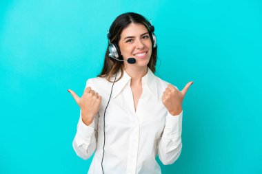 Tele pazarlamacı İtalyan kadın mavi arka planda izole edilmiş bir kulaklıkla çalışıyor. Baş parmakları havada ve gülümsüyor.