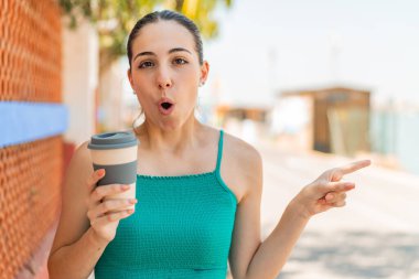 Genç ve güzel bir kadın açık havada kahve tutuyor. Şaşırmış ve işaret ediyor.