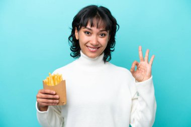 Elinde kızarmış cips tutan genç Arjantinli kadın mavi arka planda izole edilmiş parmaklarıyla tamam işareti gösteriyor.