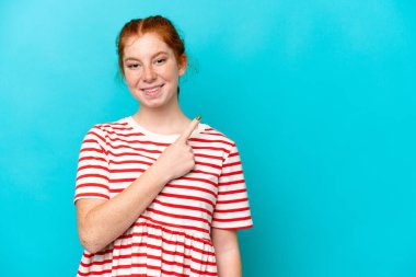 Mavi arka planda izole edilmiş genç bir kadın bir ürünü sunmak için kenarı işaret ediyor.