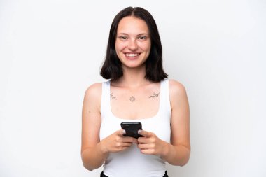 Beyaz arka planda izole edilmiş genç beyaz kadın kameraya bakıyor ve cep telefonunu kullanırken gülümsüyor.