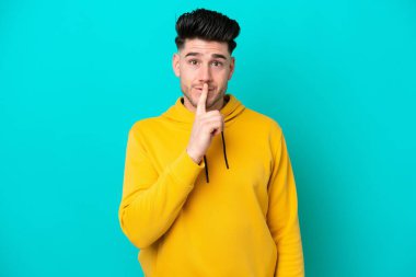 Mavi arka planda izole edilmiş genç beyaz adam sessiz bir jest işareti gösteriyor. Parmağını ağzına sokuyor.