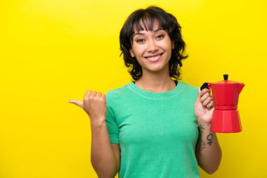 Arjantinli genç bir kadın elinde kahve makinesini sarı arka planda izole bir şekilde tutarak bir ürün sunuyor.