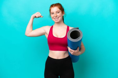 Genç spor kırmızısı kadın yoga derslerine giderken mavi arka planda izole edilmiş bir minder tutuyor güçlü bir jest yapıyor.