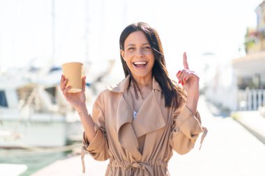 Açık havada elinde kahve tutan genç bir kadın harika bir fikri işaret ediyor.