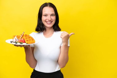 Sarı arka planda gözleme tutan genç beyaz kadın bir ürünü sunmak için yan tarafı işaret ediyor.