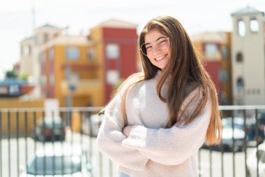 Dışarıda gözlüklü, kolları çapraz ve ileriye bakan beyaz bir kadın.