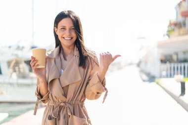 Açık havada elinde kahve tutan genç bir kadın bir ürün sunmak için yan tarafı işaret ediyor.