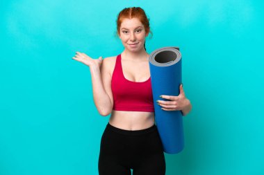 Genç spor kırmızısı kadın yoga derslerine giderken mavi arka planda izole edilmiş bir minderi tutarken ellerini kaldırırken şüpheleri oluyor.