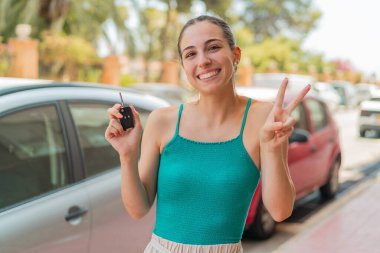 Dışarıda arabanın anahtarlarını tutan genç ve güzel bir kadın gülümsüyor ve zafer işareti gösteriyor.