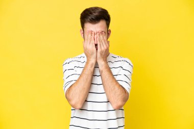 Sarı arka planda izole edilmiş genç, beyaz bir adam elleriyle gözlerini kapatıyor.