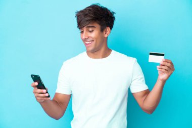 Kafkasyalı genç adam mavi arka planda izole edilmiş cep telefonuyla kredi kartıyla alışveriş yapıyor.