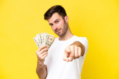Kafkasyalı genç bir adam, sarı arka planda izole edilmiş bir sürü para alıyor mutlu bir ifadeyle ön tarafı gösteriyor.