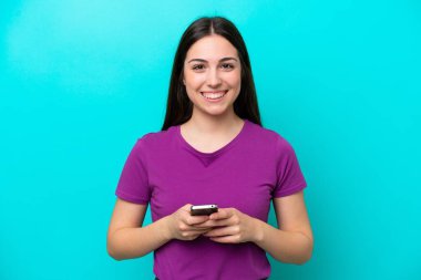 Mavi arka planda izole edilmiş genç bir kız kameraya bakıyor ve cep telefonunu kullanarak gülümsüyor.
