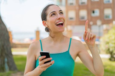 Açık havada cep telefonu kullanan genç ve güzel bir kadın çözümü bulmak için parmağını kaldırıyor.