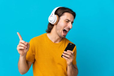 Genç ve yakışıklı bir adam mavi arka planda izole edilmiş. Cep telefonuyla müzik dinliyor ve şarkı söylüyor.