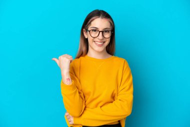 Mavi arka planda izole edilmiş genç beyaz kadın bir ürünü sunmak için kenarı işaret ediyor.