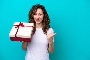 Elinde mavi arka planda izole edilmiş bir hediye tutan beyaz bir kadın bir ürünü sunmak için yan tarafı işaret ediyor.