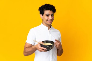 Genç Venezuelalı bir adam sarı arka planda izole edilmiş ve elinde çubuklarla bir kase erişte tutarken onu işaret ediyor.