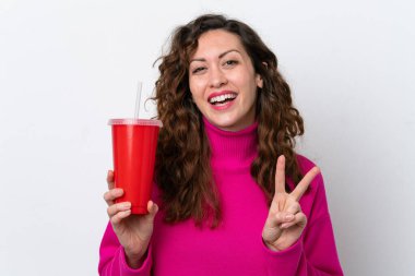 Beyaz arka planda izole edilmiş gazoz içen beyaz kadın gülümsüyor ve zafer işareti gösteriyor.