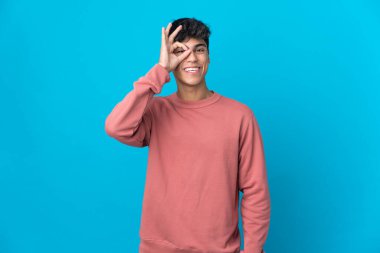 İzole edilmiş mavi arka planda genç bir adam parmaklarıyla tamam işareti gösteriyor.