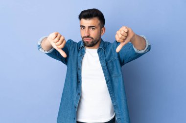 İzole edilmiş mavi arka planda beyaz bir adam baş parmağını iki eliyle aşağı gösteriyor.