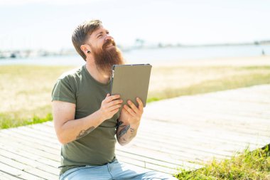 Kızıl saçlı, sakallı bir adam elinde tabletle dışarı bakıp gülümsüyor.