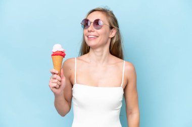 Mavi arka planda izole edilmiş dondurma tutan mayolu genç sarışın kadın gülümserken yukarı bakıyor.