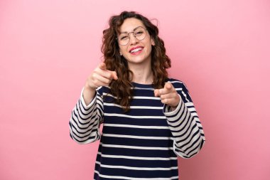 Pembe arka planda izole edilmiş genç beyaz kadın mutlu bir ifadeyle ön tarafı işaret ediyor.