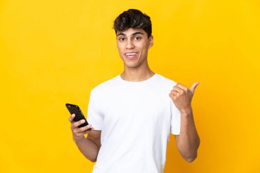 Genç adam sarı arka planda cep telefonu kullanıyor ve yan tarafı işaret ediyor.