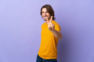 Mor arka planda izole edilmiş genç yakışıklı gülümseyen ve zafer işareti gösteren