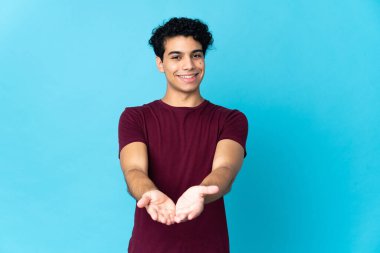 Genç Venezuelalı bir adam mavi arka planda izole edilmiş bir şekilde bir ilan vermek için fotokopi uzayı avucunun içinde tutuyor.