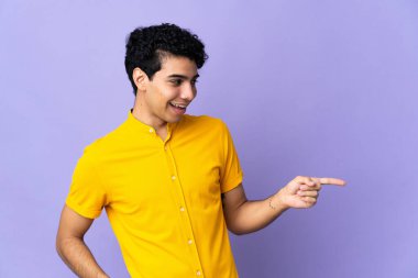 Genç Venezuelalı bir adam mor arka planda izole edilmiş bir şekilde parmak ucunda bir ürün sunuyor.