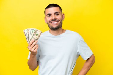 Kafkasyalı genç adam çok para alıyor. Sarı arka planda izole edilmiş. Kolları kalçasında ve gülümseyerek poz veriyor.