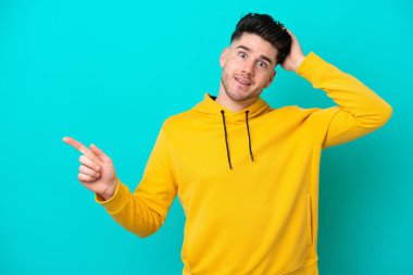 Kafkasyalı genç adam mavi arka planda izole edildi. Şaşırdı ve işaret etti.